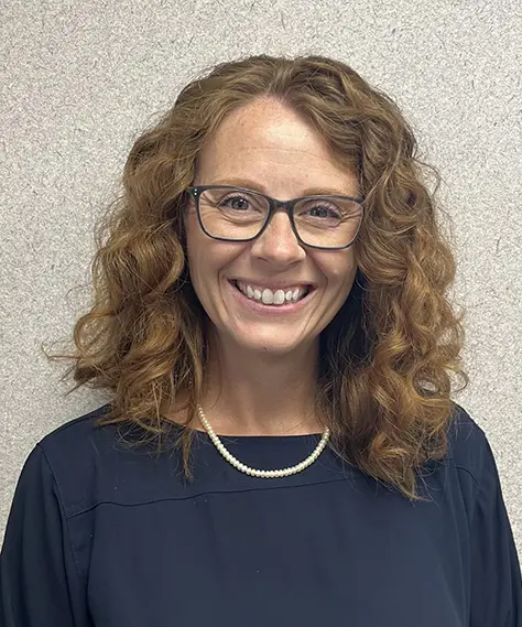 A woman with curly red hair, glasses, and a pearl necklace smiles at the camera. She is wearing a dark blouse and standing in front of a light-colored textured wall.