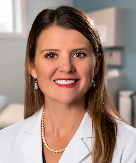 A woman with long brown hair, wearing a white lab coat, pearl necklace, and pearl earrings, smiles in a medical office setting.